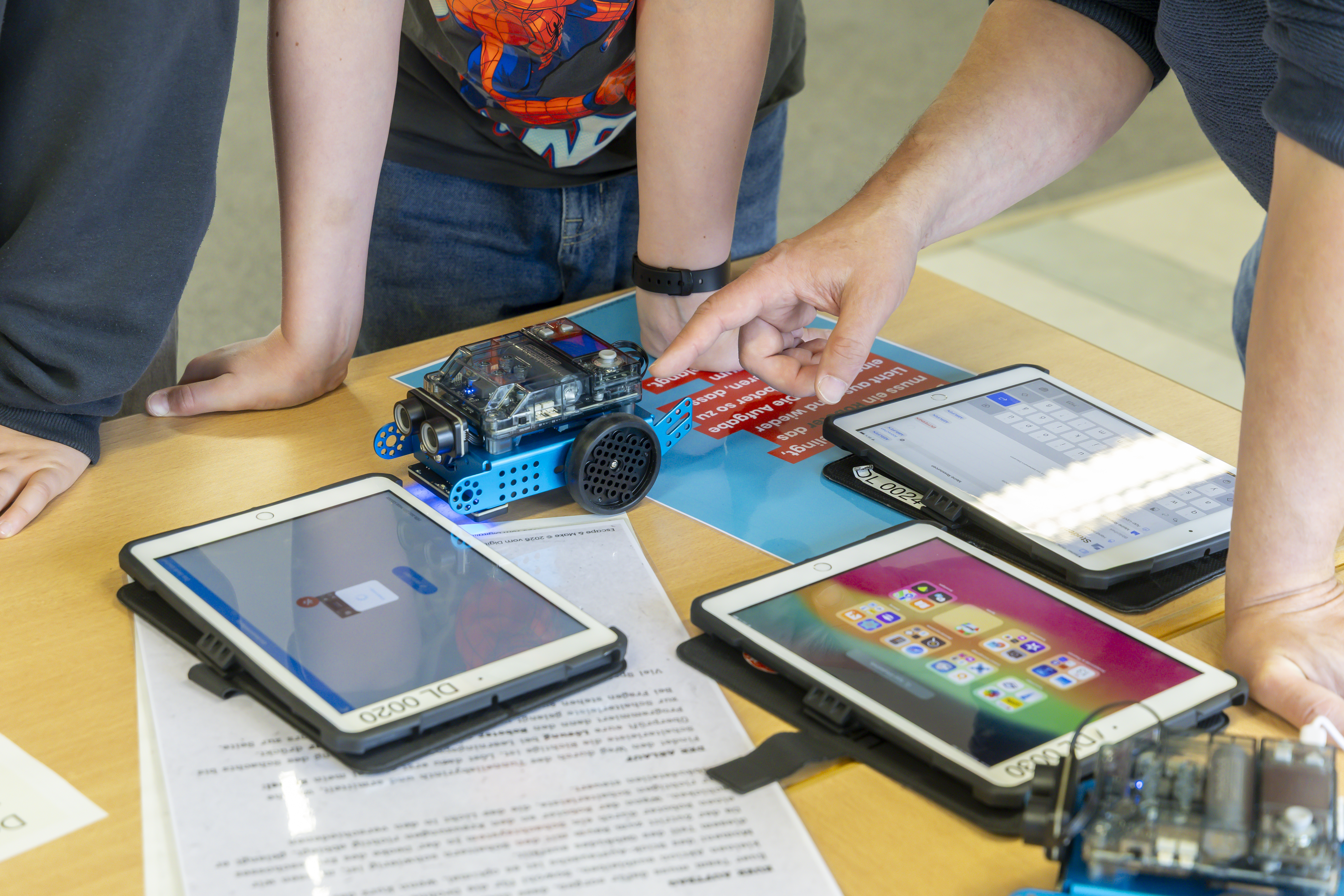 Zwei Tablets liegen auf einem Tisch. Drei Kinder stehen daneben, eins zeigt auf deinen Bildschirm