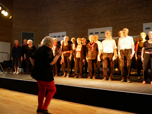 Ein Chor steht auf der Bühne und singt. Die Chorleiterin steht vor der Gruppe.
