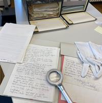 Auf einem Tisch liegen zwei geöffnete Archivkisten sowie mehrere Unterlagen mit handschriftlichen Notizen, weiße Handschuhe und eine Lupe zur Untersuchung des Archivmaterials