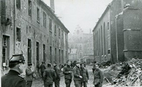 Schwarz-weiß-Foto: Eine Gruppe Männer vor einem Trümmergrundstück in einer Straße, deren Häuser deutliche Kriegsschäden haben