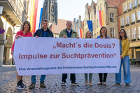 Menschen des Arbeitskreises stehen auf dem Prinzipalmarkt und halten ein Plakat in die Kamera, auf dem steht: "Macht's die Dosis? Impulse zur Suchtprävention."