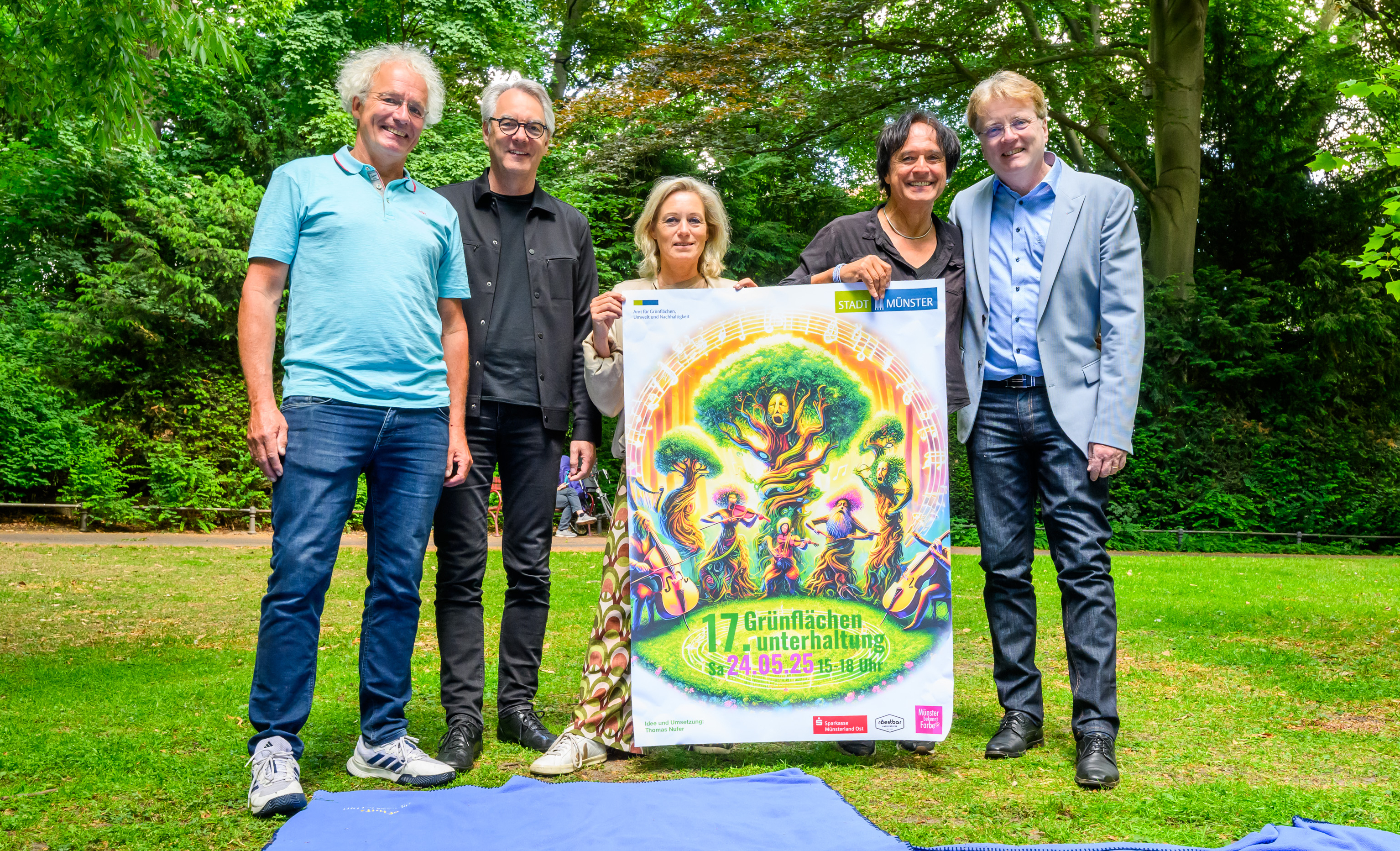 Fünf Personen stehen auf einer Wiese hinter einer Picknickdecke und zeigen ein Plakat