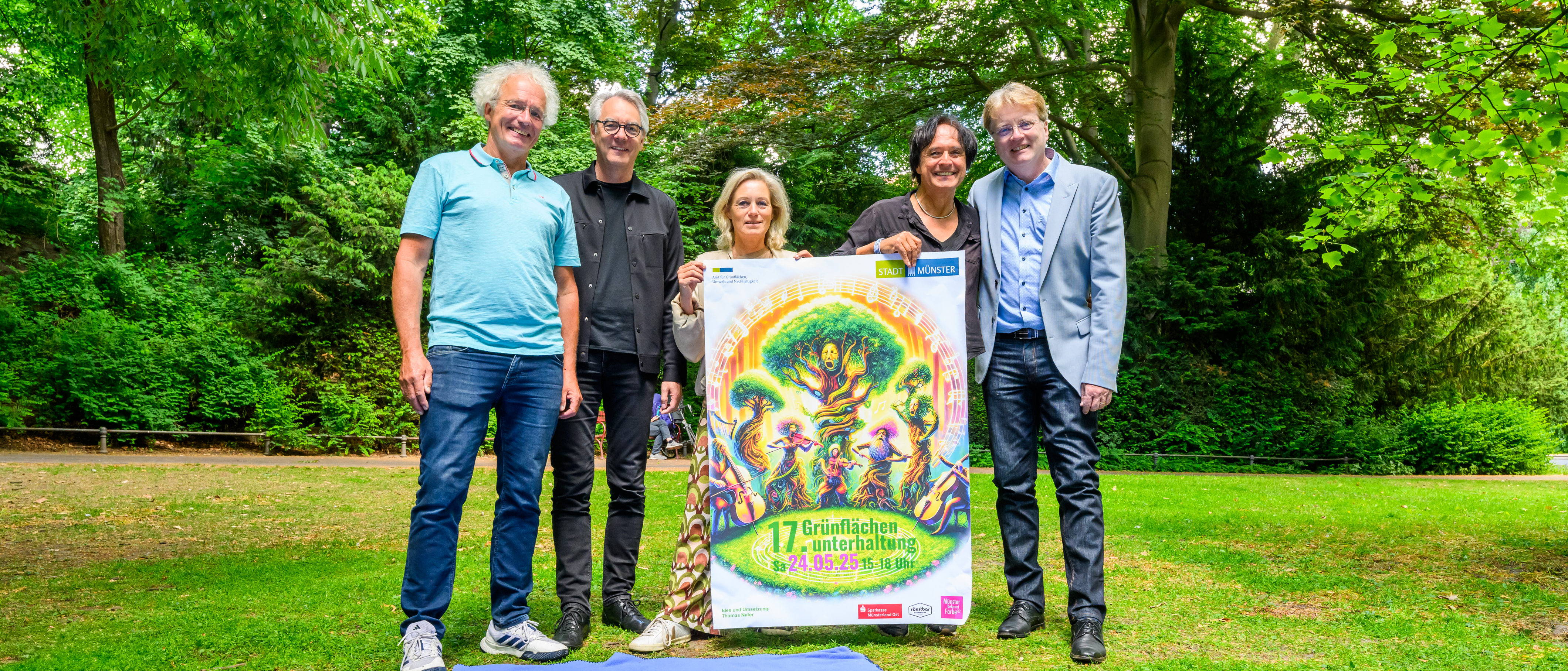 Fünf Personen stehen auf einer Wiese hinter einer Picknickdecke und zeigen ein Plakat