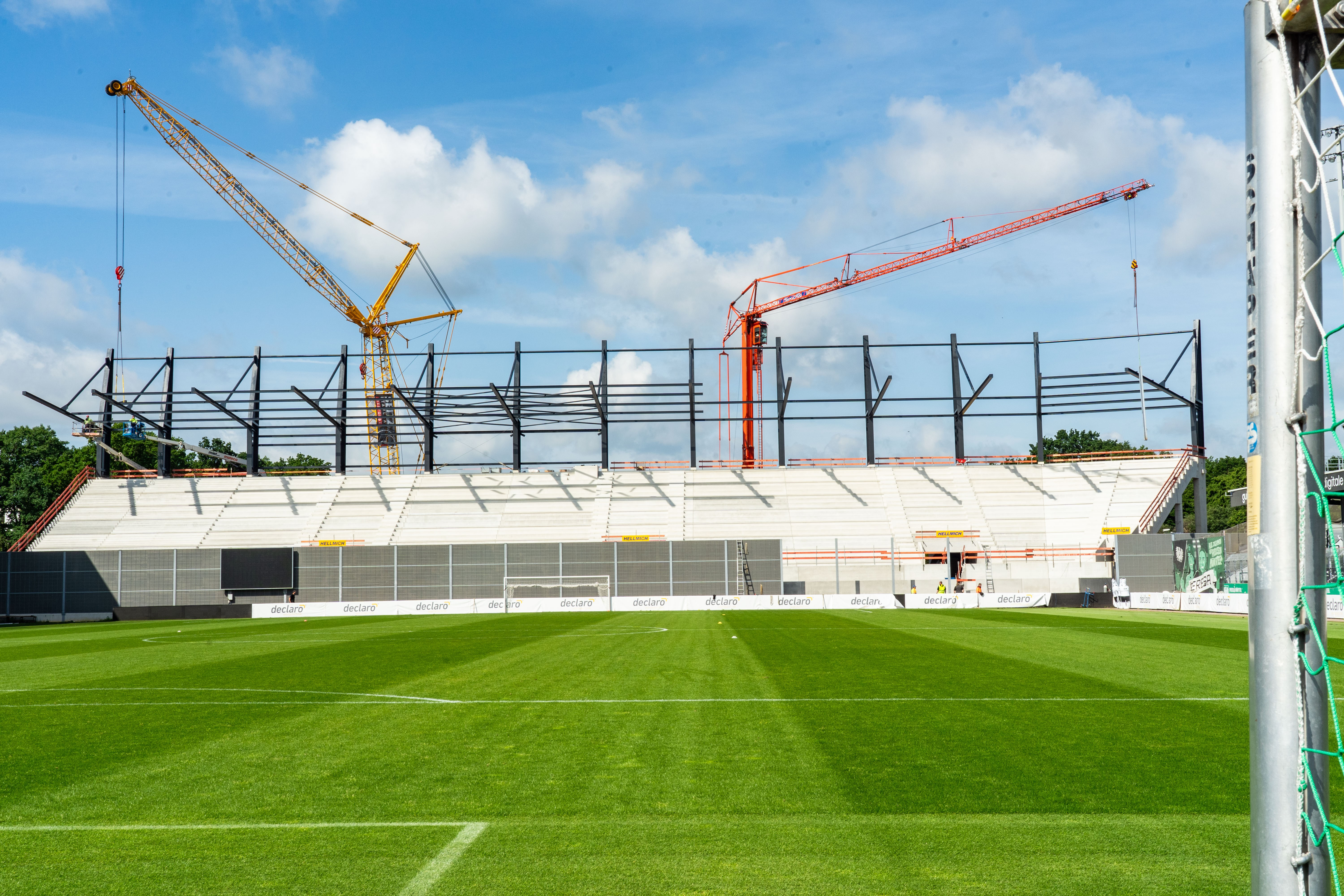Ein frontaler Blick auf die Westtribüne, die noch nicht fertiggestellt ist. Im Vordergrund das Fußballfeld, hinter der Tribüne ragen zwei Baukräne hoch.