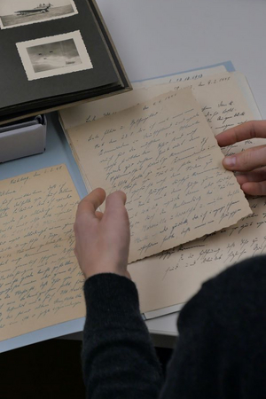 Auf einem Tisch liegen alte Zettel mit Handschrift. Zwei Hände halten einen Zettel fest.