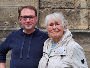 links ein Mann mit Brille und blauem Pullover, rechts eine Frau mit weißen Haaren, Brille und einer weißen Jacke. Auf der Jacke ist ein Button mit der Aufschrift "Omas gegen Rechts"