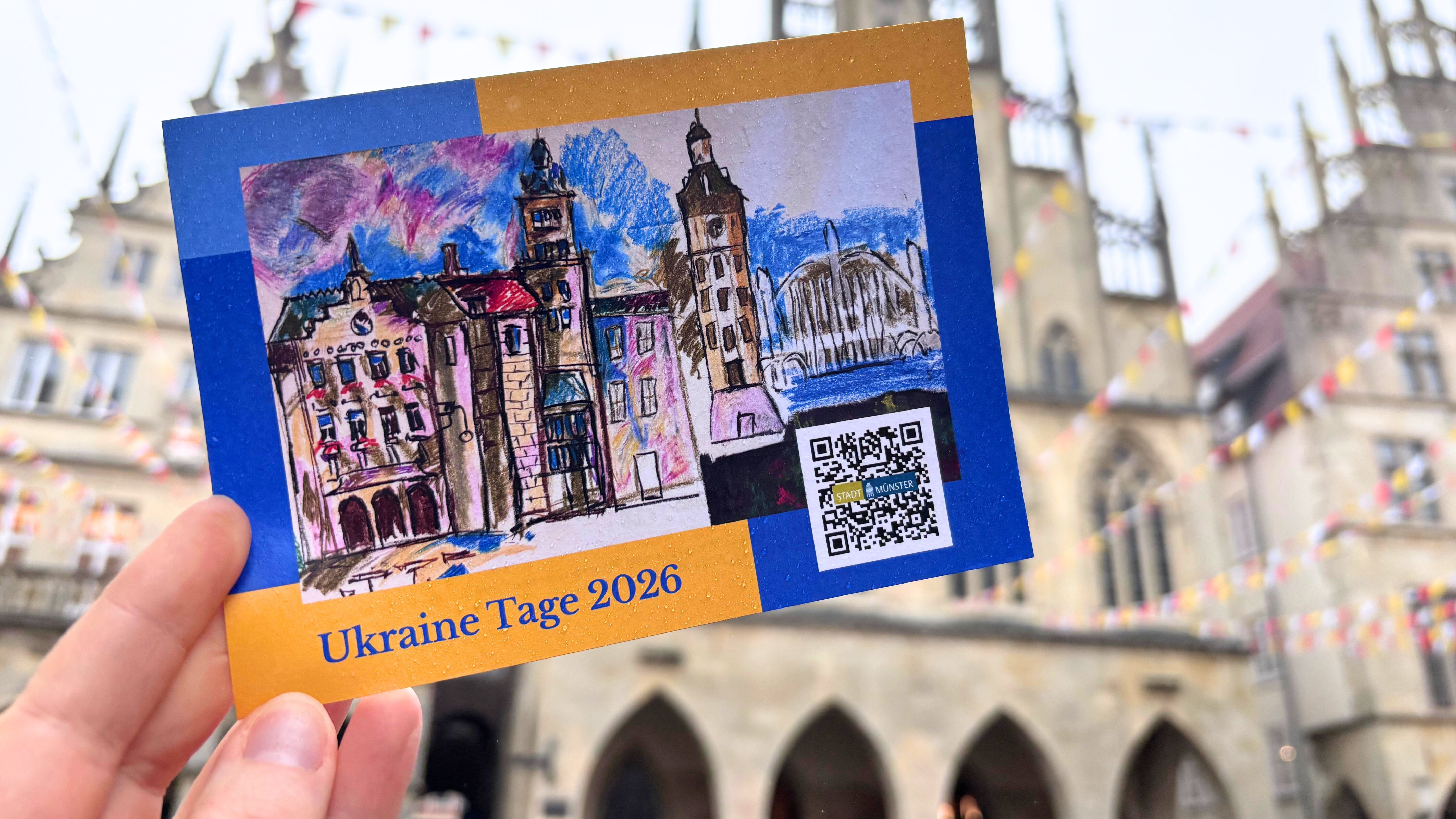 Eine Hand hält eine Postkarte zu der Veranstaltungsreihe Ukraine-Tage hoch. Im Hintergrund ist das Rathaus am Prinzipalmarkt unscharf zu erkennen. 
