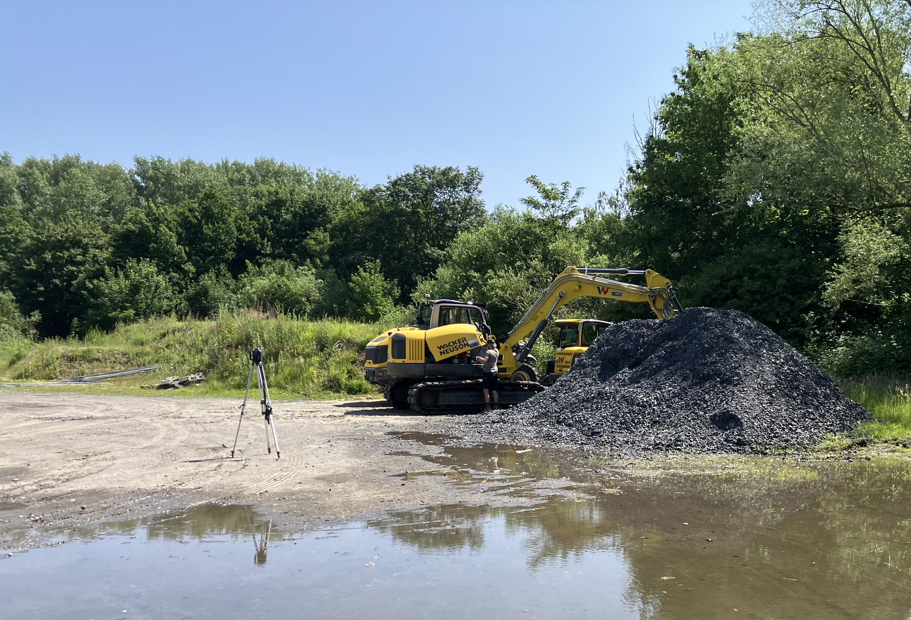 Ein Bagger steht vor Bäumen. Im Vordergrund ist Wasser.