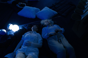 Zwei Performerinnen liegen in einem dunkelblauen Zelt mit den Köpfen nebeneinander. Links von ihnen leuchtet eine runde Lampe.