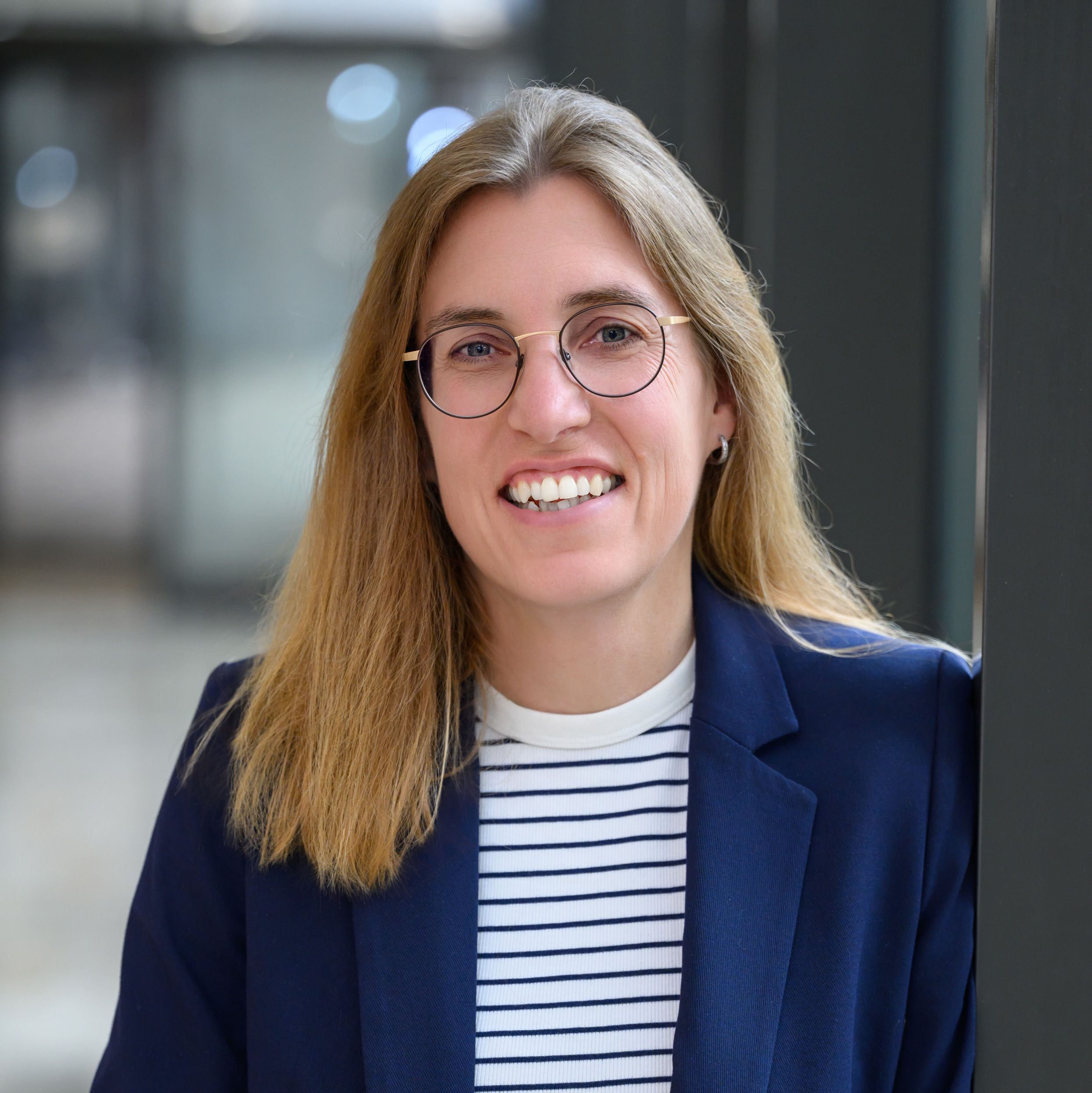 Eine Frau mit braunen Haaren, Brille, gestreiftem Shirt und Blazer
