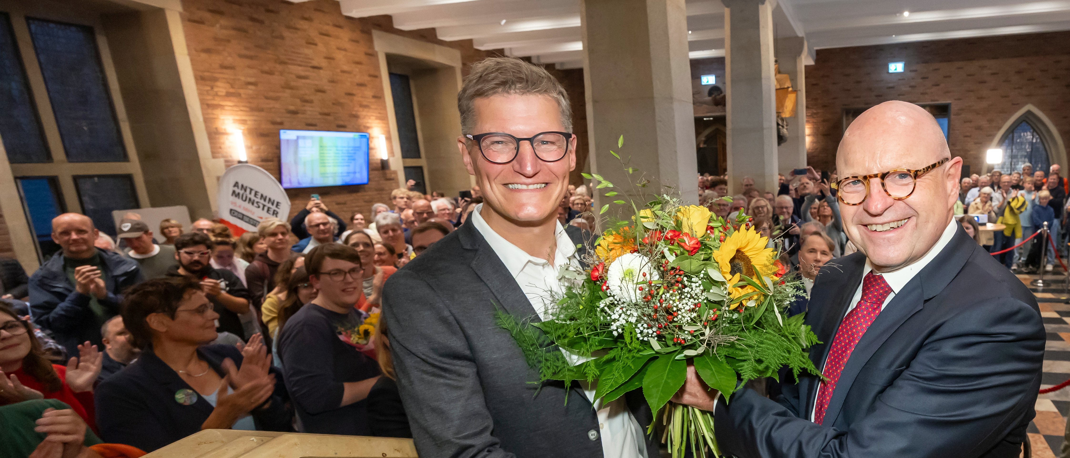 Tilman Fuchs und Markus Lewe mit Blumenstrauß in der gut besuchten Bürgerhalle im Historischen Rathaus in Münster