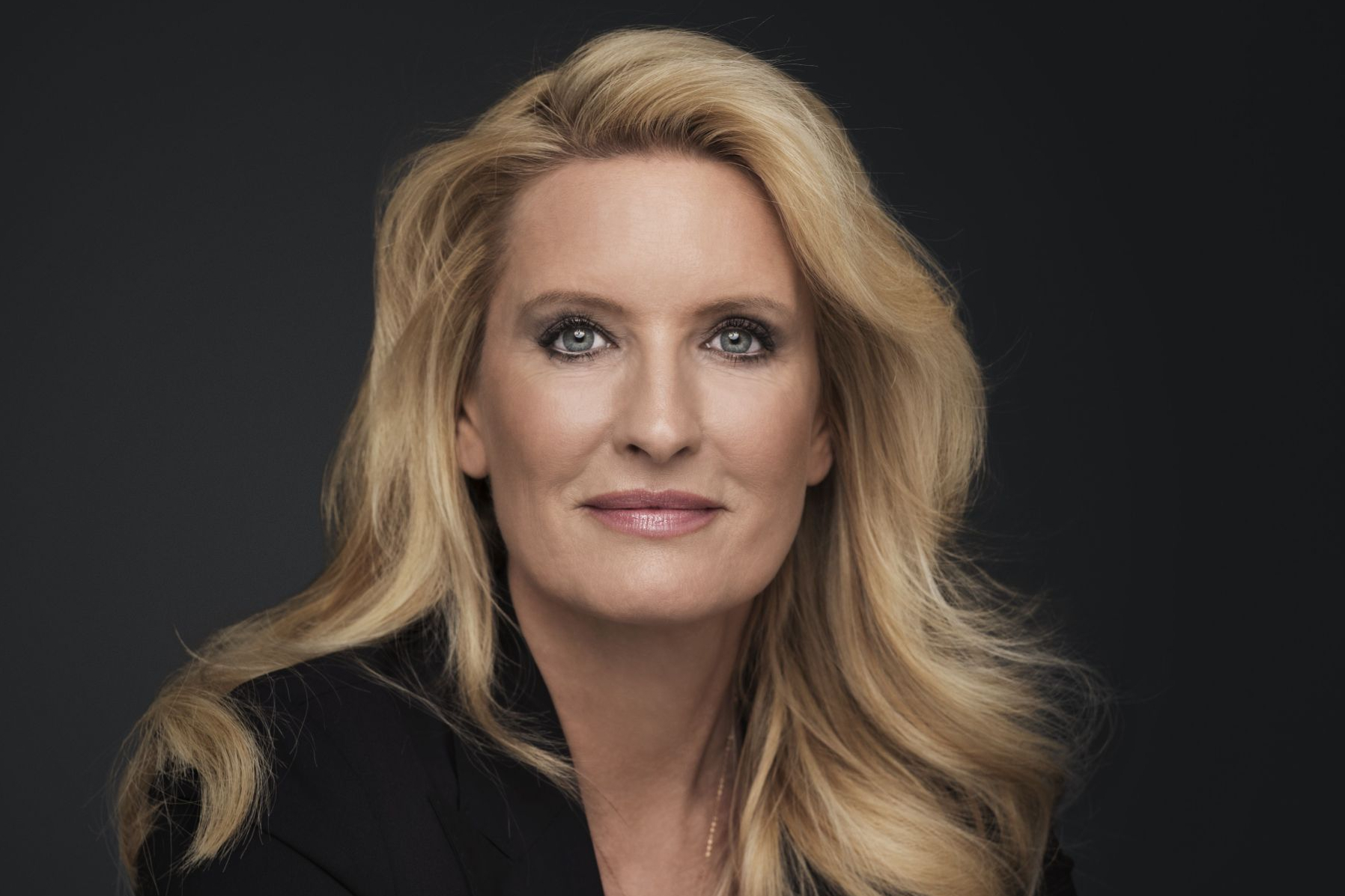 Frau mit langen blonden Haaren, schwarzem Blazer und weißem Oberteil vor dunklem Hintergrund