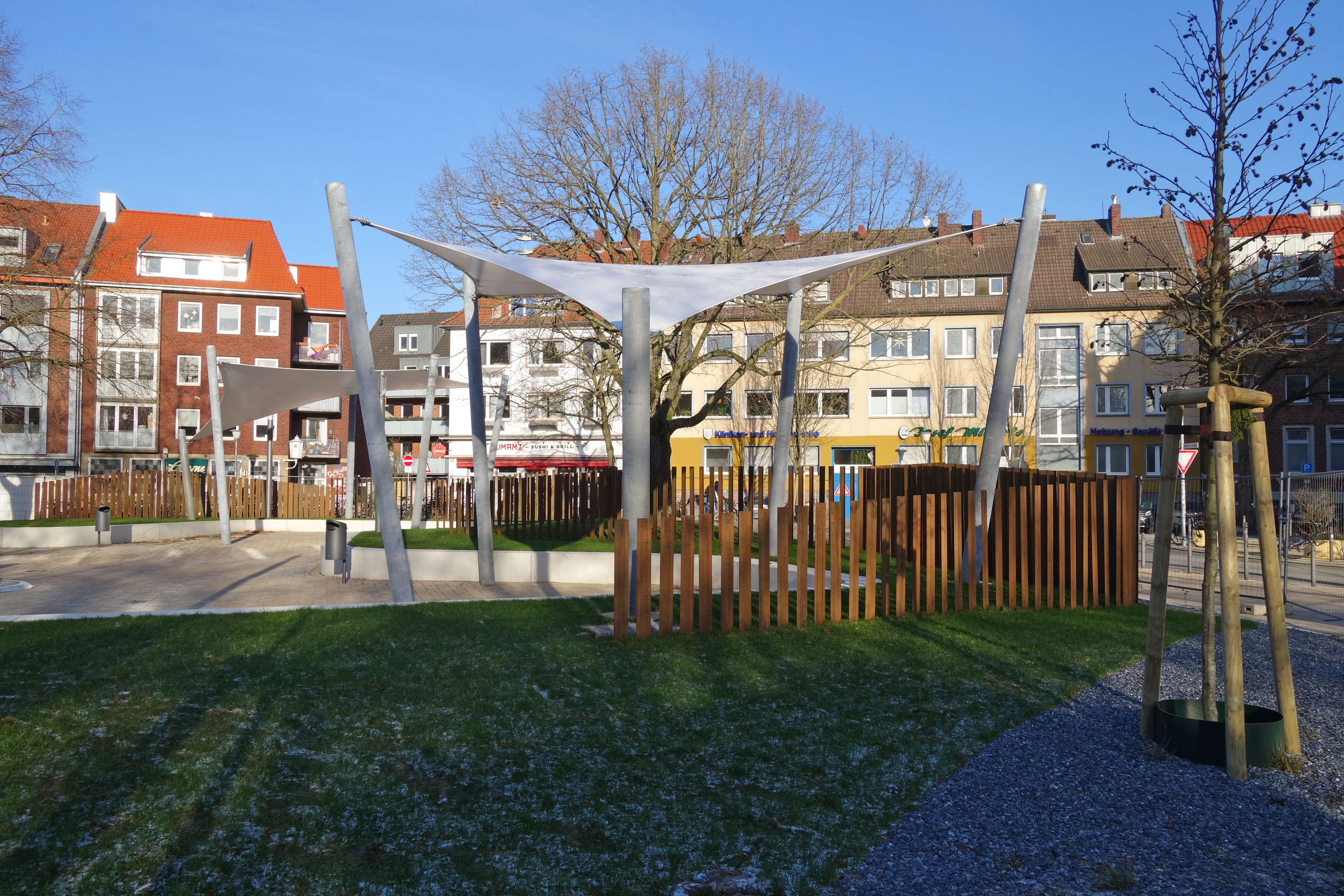 Blick auf den Bremer Platz mit zwei Segelkonstruktionen, einem Holzzaun, Wiese und einem asphaltierten Platz. Im Hintergrund mehrstöckige Häuser.