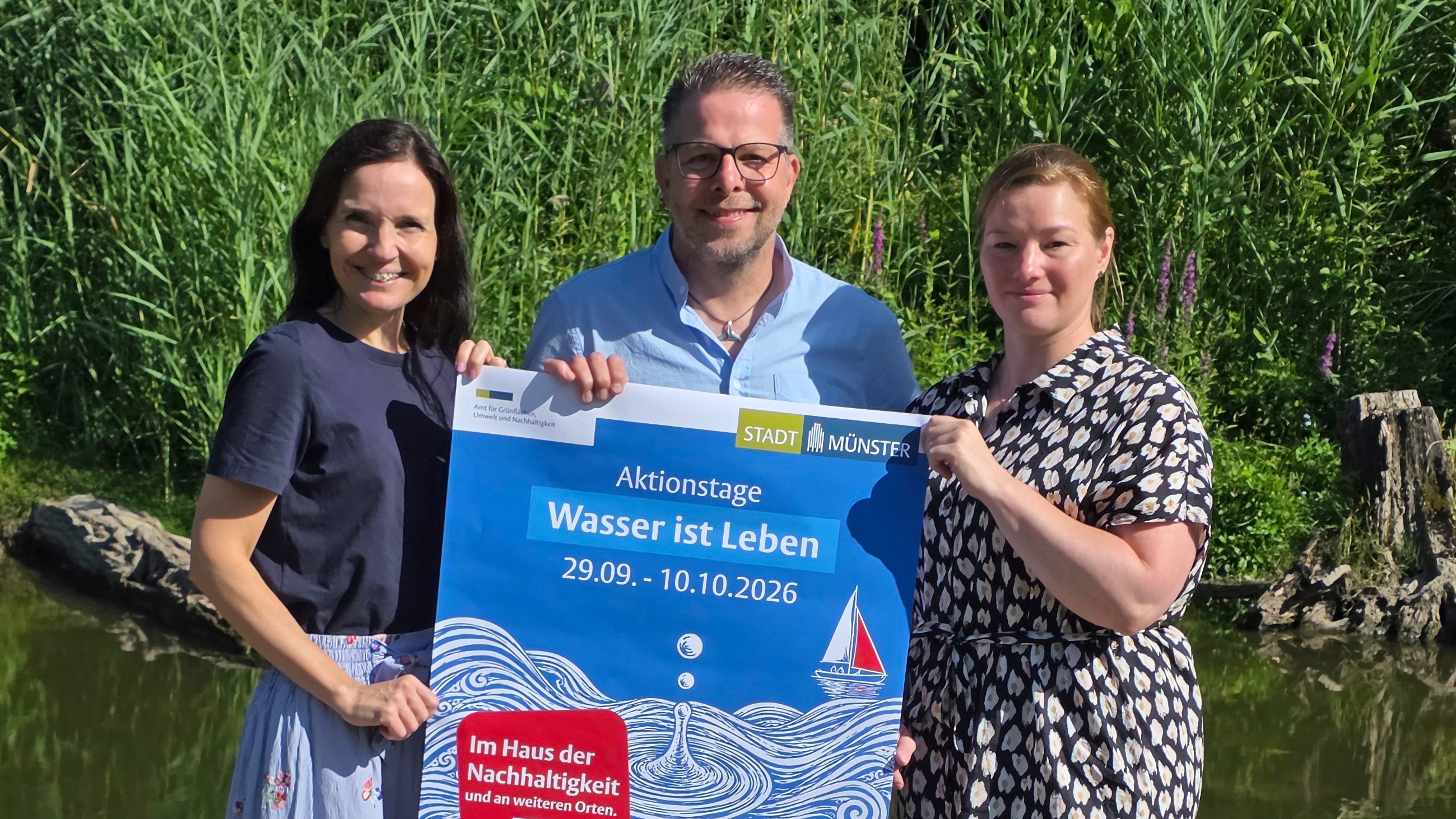 Das Foto zeigt die projektverantwortlichen Mitarbeitenden der Stadt Münster Julia Ebert, Sven Kentrup und Jessica Fischer, die das Plakat der Aktionstage 2026 halten, im Hintergrund Schilfgras und ein Teich. Das Poster zeigt ein Segelboot, Wellen, Wassertropfen auf blauem Grund mit roten Akzenten, Schriftzug "Aktionstage Wasser ist Leben", 29.09.-10.10.2026.