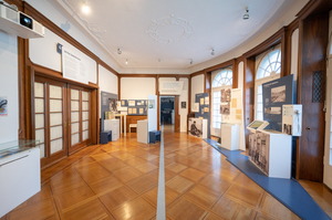 Ein großer Raum in einem herrschaftlichen Haus mit Parkettboden. An den Wänden sind Fotos und Texte zu sehen.