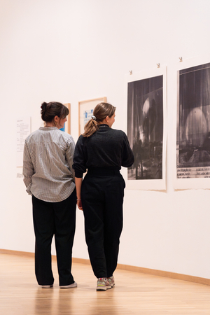 Zwei Frauen stehen im Ausstellungsraum und betrachten die Werke an der Wand.
