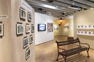 Eine historische Parkbank steht im Ausstellungsraum. An den Wänden zahlreiche Postkarten und Mit-mach-Stationen.