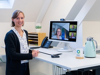 Ramona Leusch von der KLENKO steht vor einem Computer-Bildschirm, zu sehen ist eine geöffnete Zoom-Konferenz.