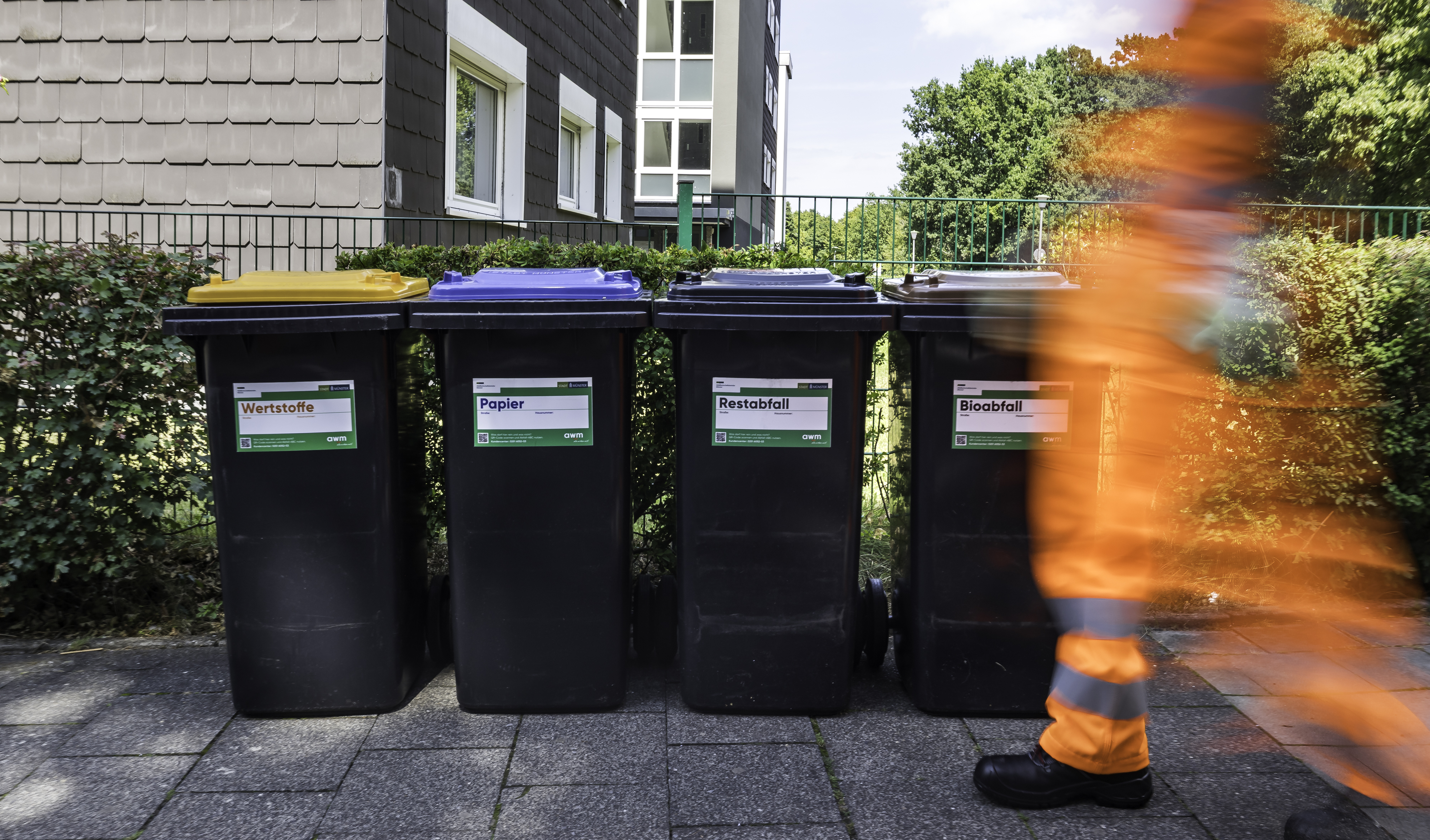 Vier Mülltonnen vor einem eingezäunten Grundstück, im Vordergrund ein schemenhafter Mitarbeiter der awm