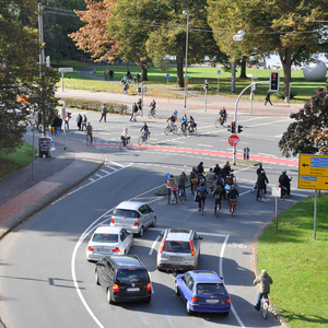 Mehrere Fahrradfahrerinnen und Fahrradfahrer, sowie einige Autos warten auf einer Straße vor einer Ampel an einer Kreuzung am Aasee während Fußgängerinnen und Fußgänger die Straße überqueren. 