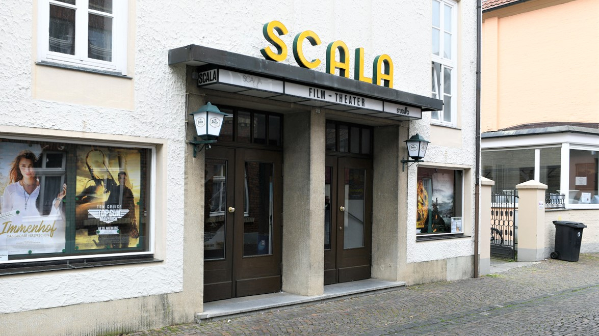 Außenansicht des Filmtheaters: Eine weiß, graue Fassade mit einer doppelten schweren Tür, über der Tür sind neongelbe Buchstaben angebracht, die den Namen Scala ergeben. Links und rechts neben der Tür sind Schaufenster mit Kinoplakaten.