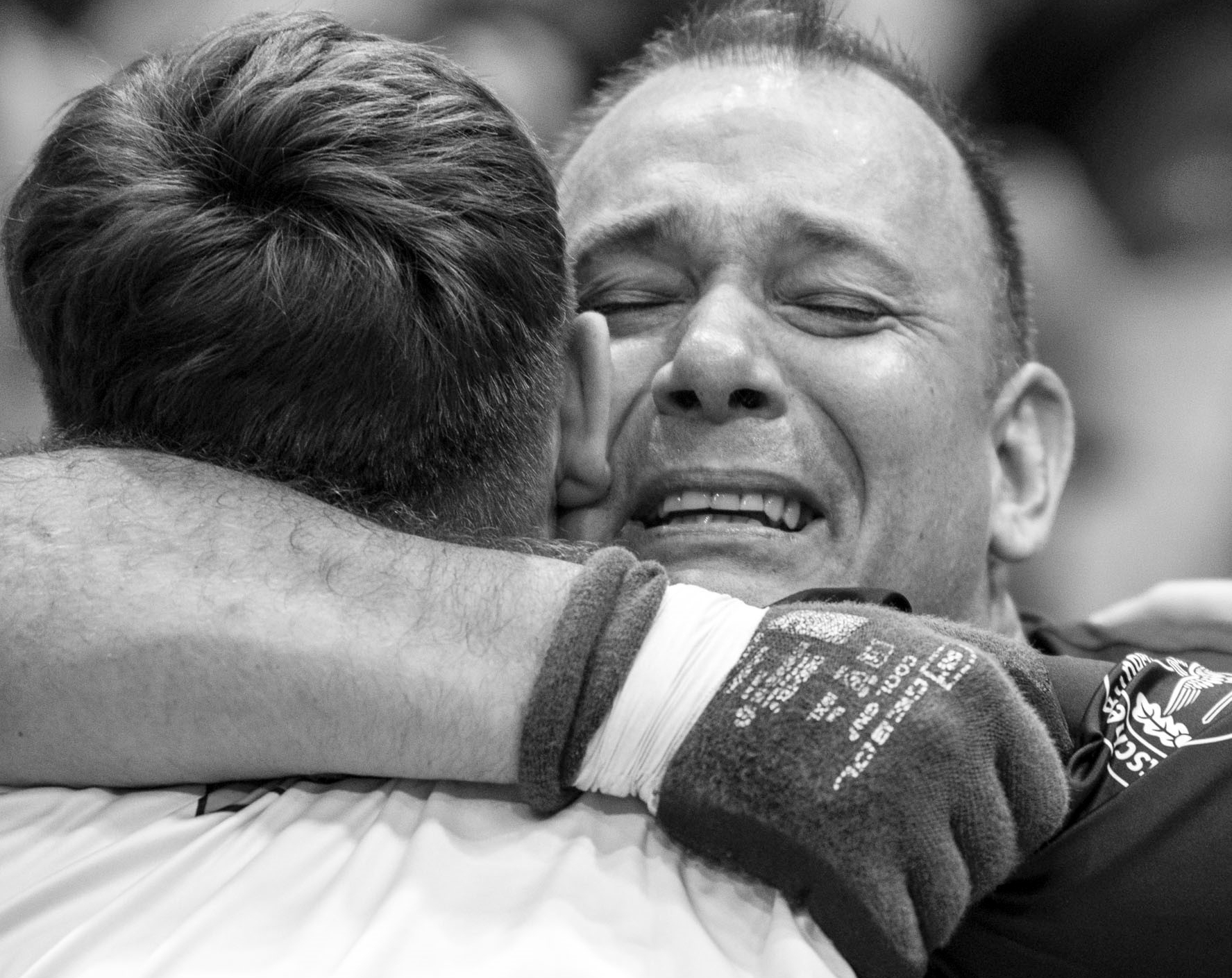 Zwei Sportler (anscheinend im Rollstuhl) umarmen sich. Der Sportler, der dem Betrachter zugewandt ist, hat Tränen in den zugekniffenen Augen, schaut aber glücklich und erleichtert aus.