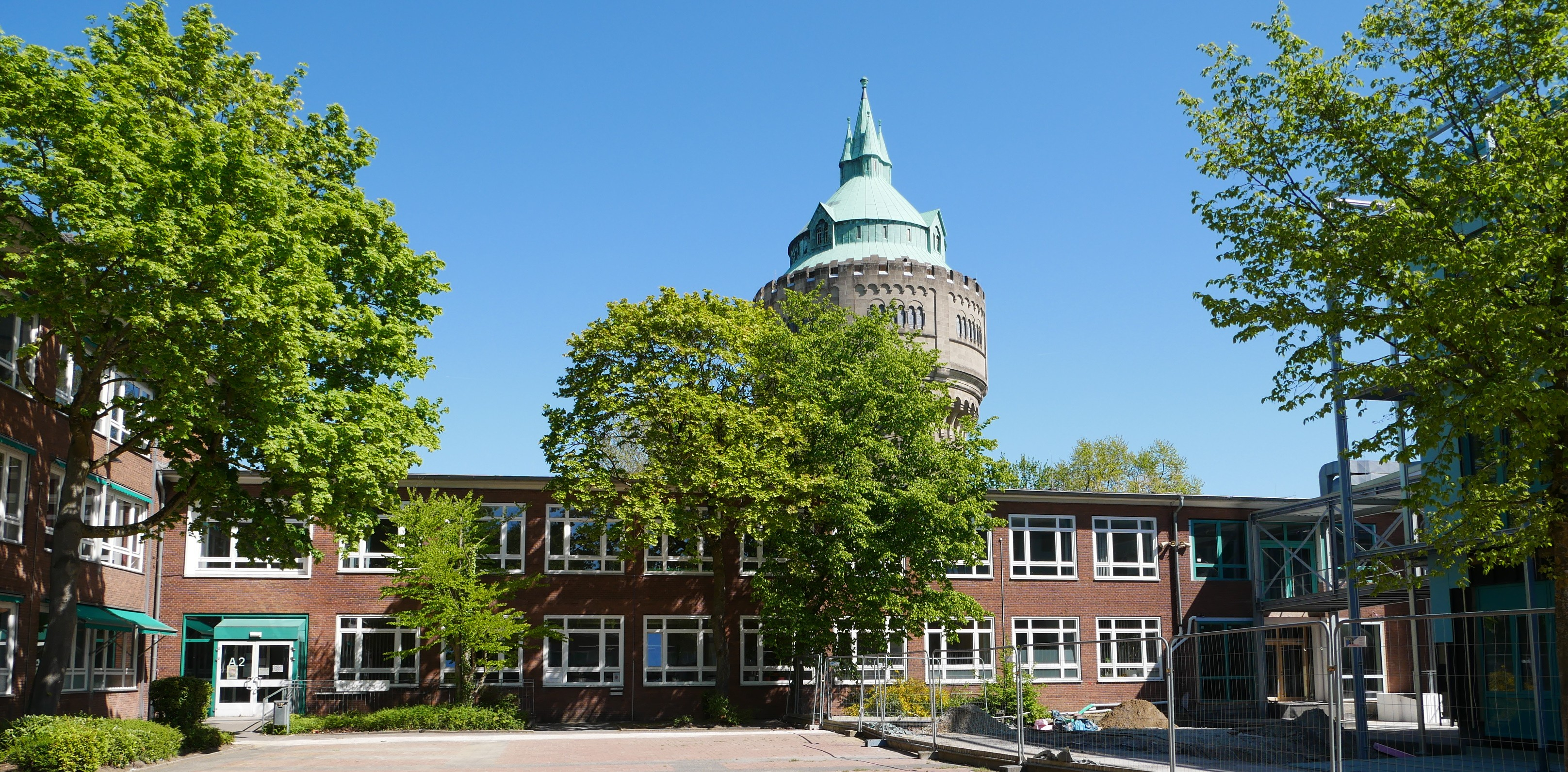 Ein Foto eines Schulgebäudes von außen, im Hintergrund ein Turm und vor dem Gebäude Hecken und Bäume