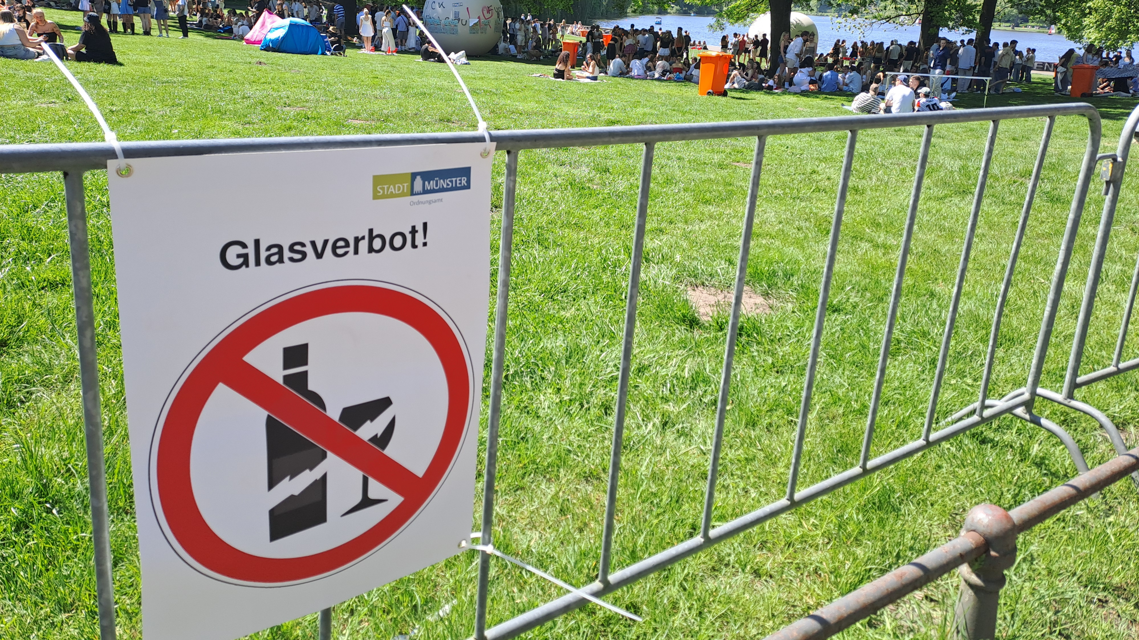 Ein Glasverbotsschild an einem Bauzaun, im Hintergrund die Aaseekugeln und der Aasee