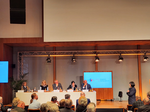 Blick aus dem Zuschauerraum auf eine Bühne, auf der an einem langen Tisch fünf Personen sitzen. Im Hintergrund ist ein Monitor, rechts steht eine Frau mit einem Mikrofon.
