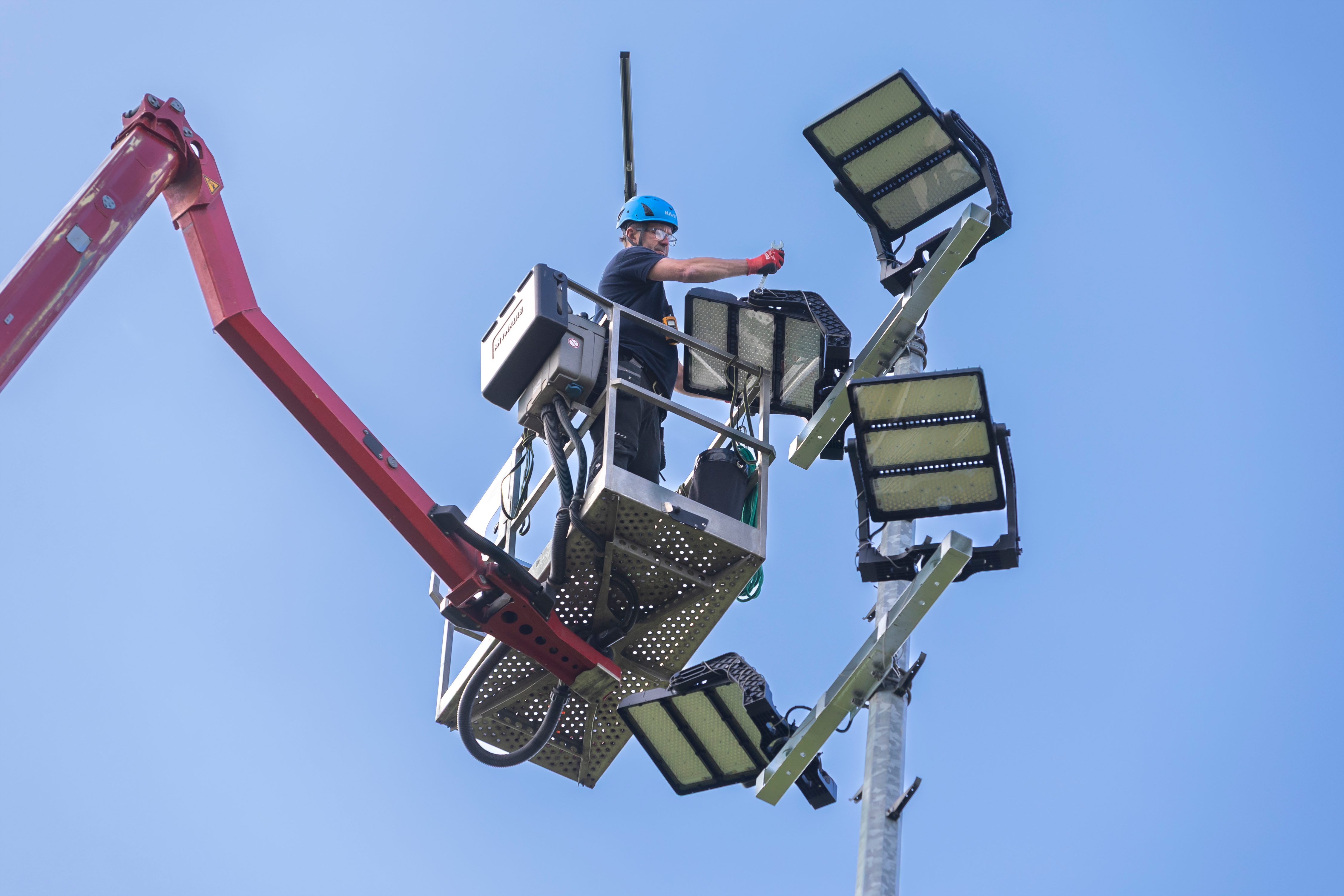 Im Korb eines Hubwagens arbeitet eine Person an einem Flutlicht, im Hintergrund blauer Himmel.