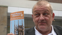TV-Ermittler Wilsberg hält die Broschüre "StattReisen Münster" ins Bild.
