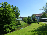 Die münstersche Aa in der Innenstadt nahe der Uni-Bibliothek. Der kleine Fluss - die münsterische Aa - fließt durch die Innenstadt. Links davon eine Wiese, auf der sich Personen ausruhen und sitzen.
