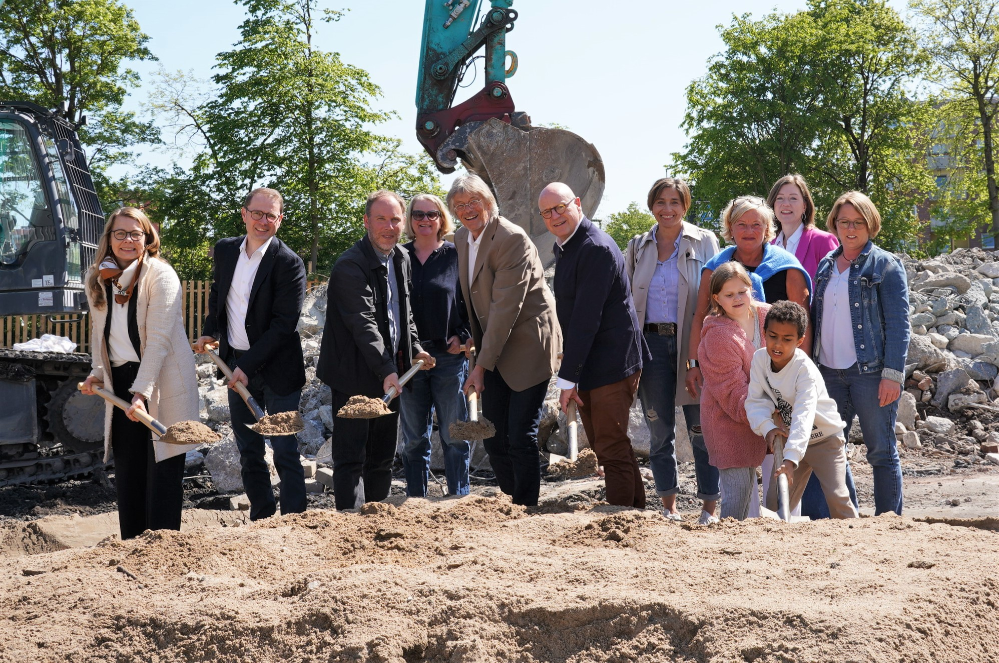 Personen stehen vor einem Bagger und halten zum teil mit Sand gefüllte Spaten in der Hand.