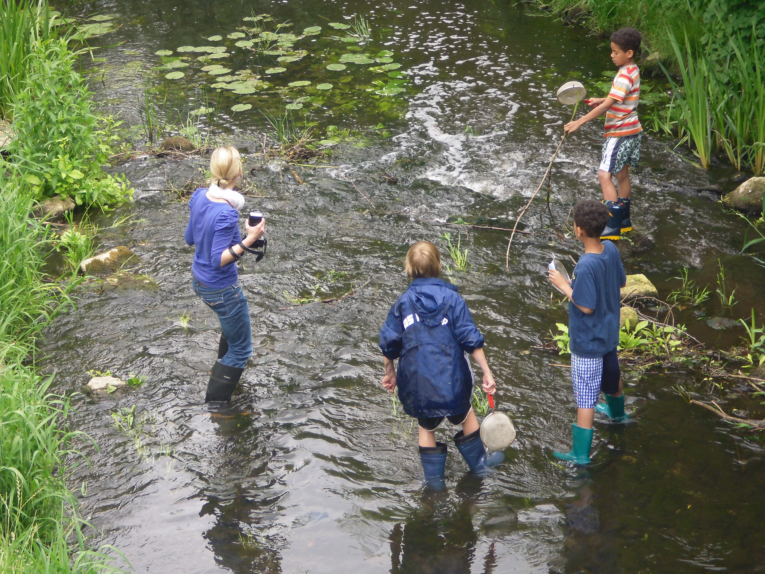 Vier ältere Kinder stehen mit Gummistiefeln in einem etwa drei Meter breiten Bachlauf.