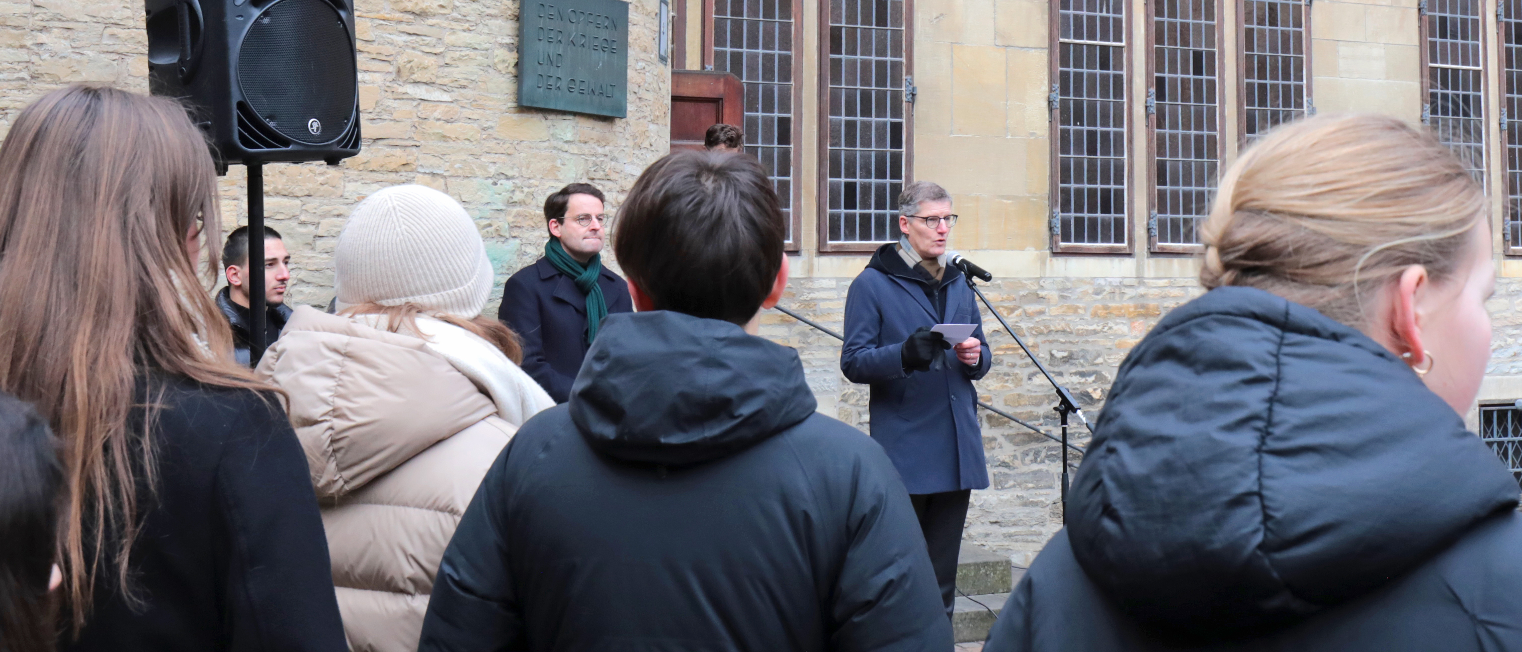 Im Vordergrund Personen, die dem Oberbürgermeister am Mikrofon im Bildhintergrund zuhören.