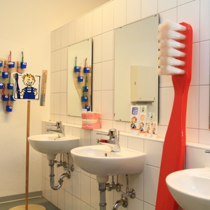 Blick in ein Kita- oder Schulbadezimmer mit drei Waschbecken und einem menschengroßen Modell einer roten Zahnbürste