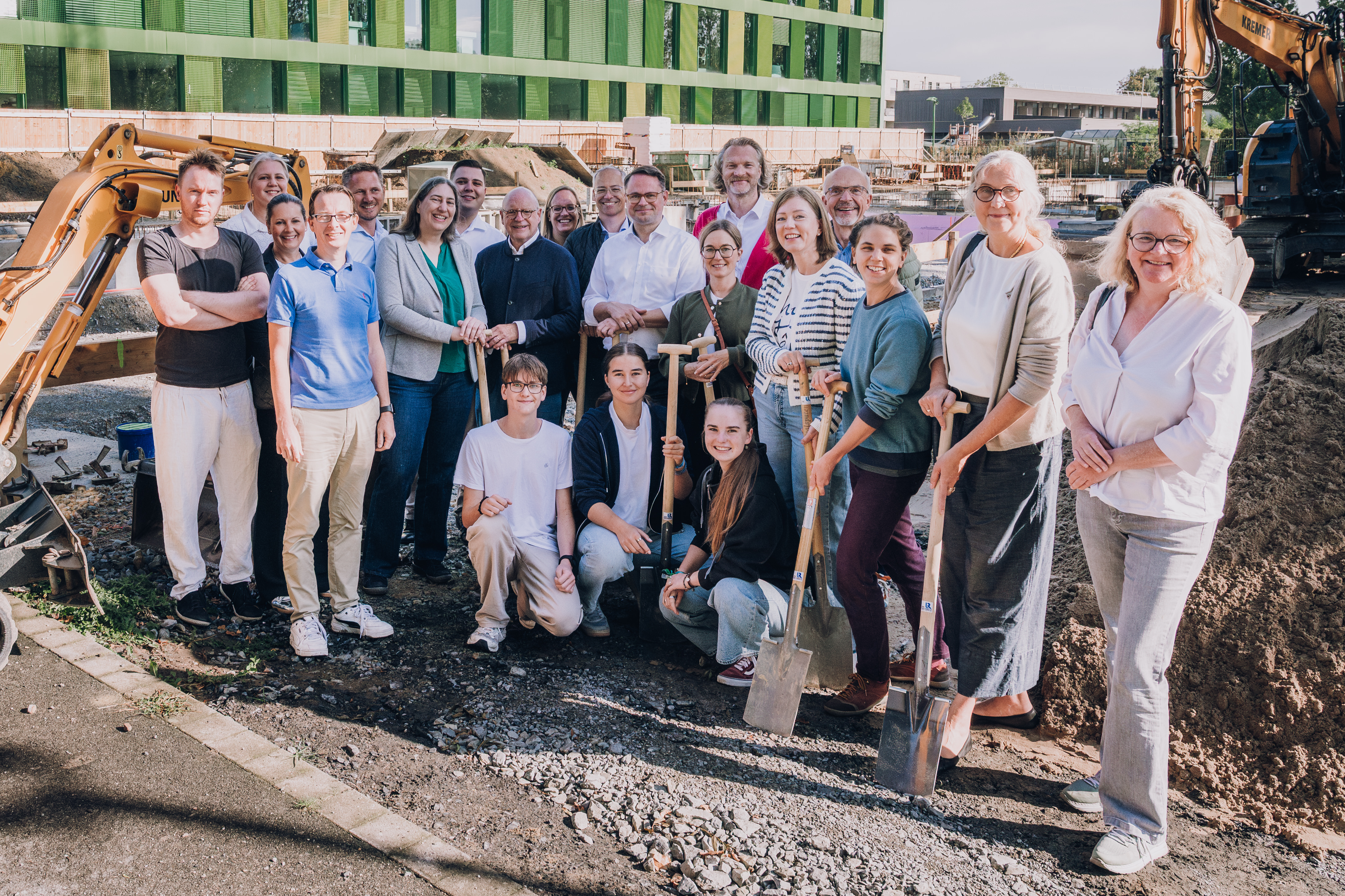 Gruppenfoto vor dem grünen Schulgebäude, links neben der Gruppe steht ein kleiner Bagger.