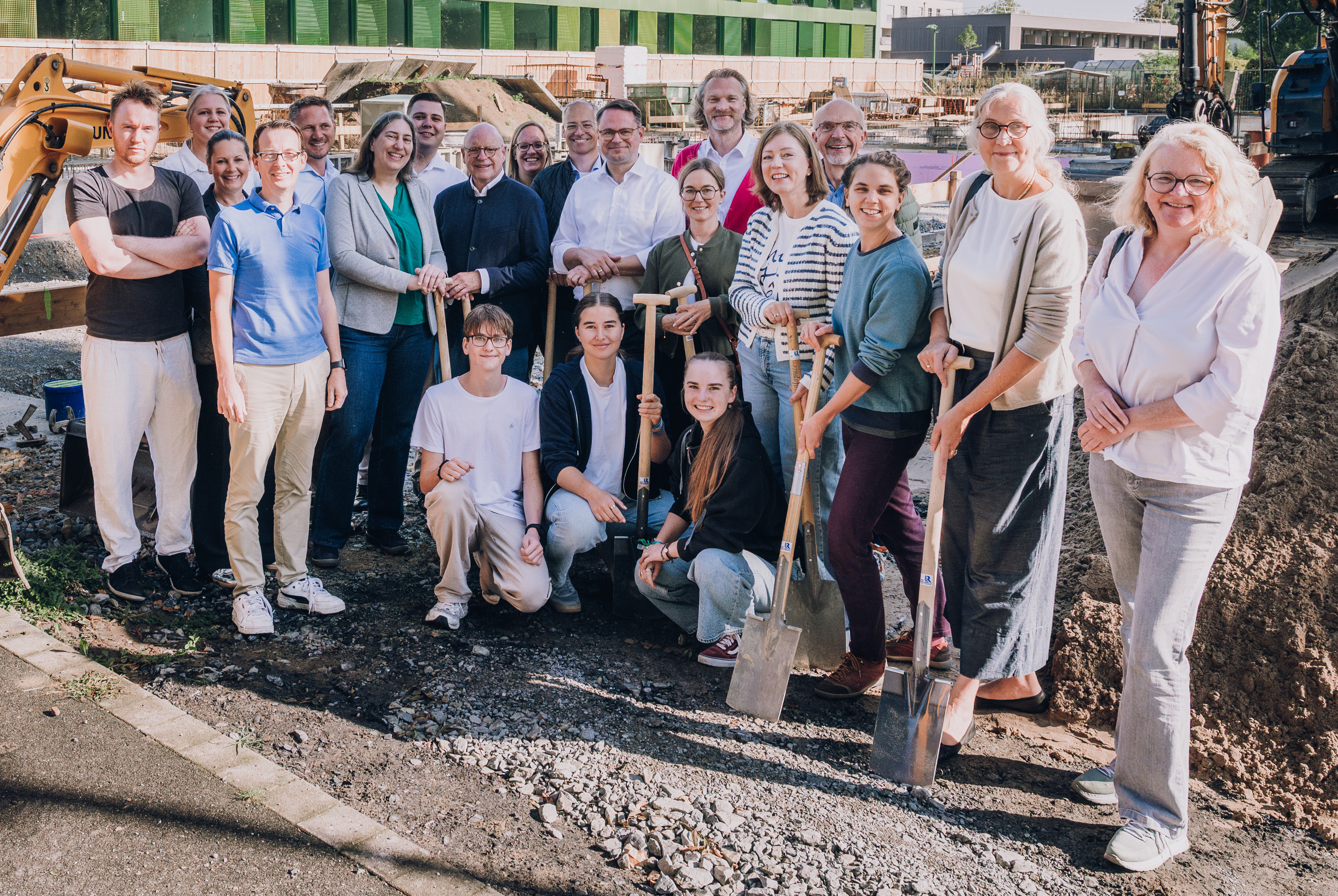 Gruppenfoto vor dem grünen Schulgebäude, links neben der Gruppe steht ein kleiner Bagger.