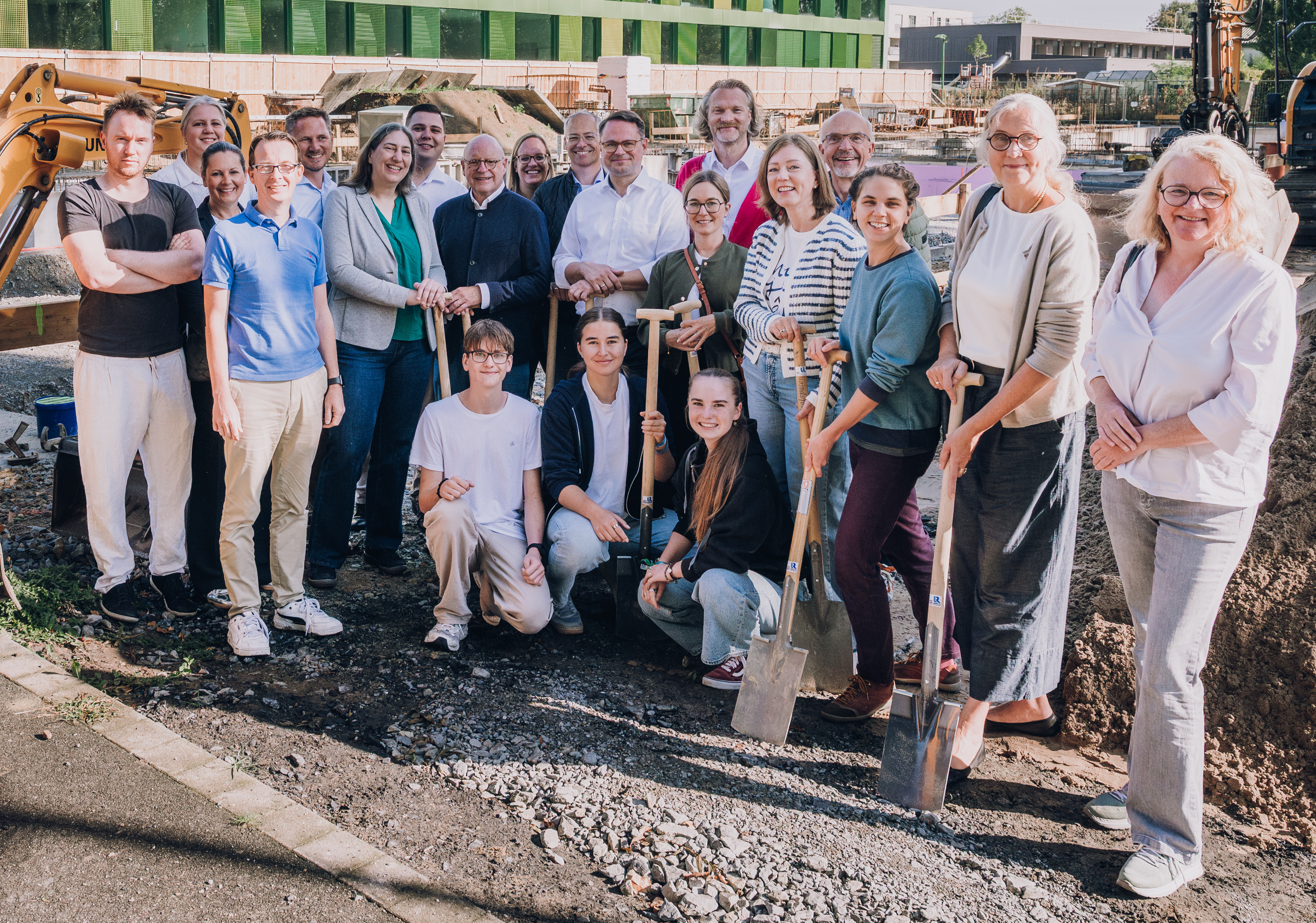 Gruppenfoto vor dem grünen Schulgebäude, links neben der Gruppe steht ein kleiner Bagger.