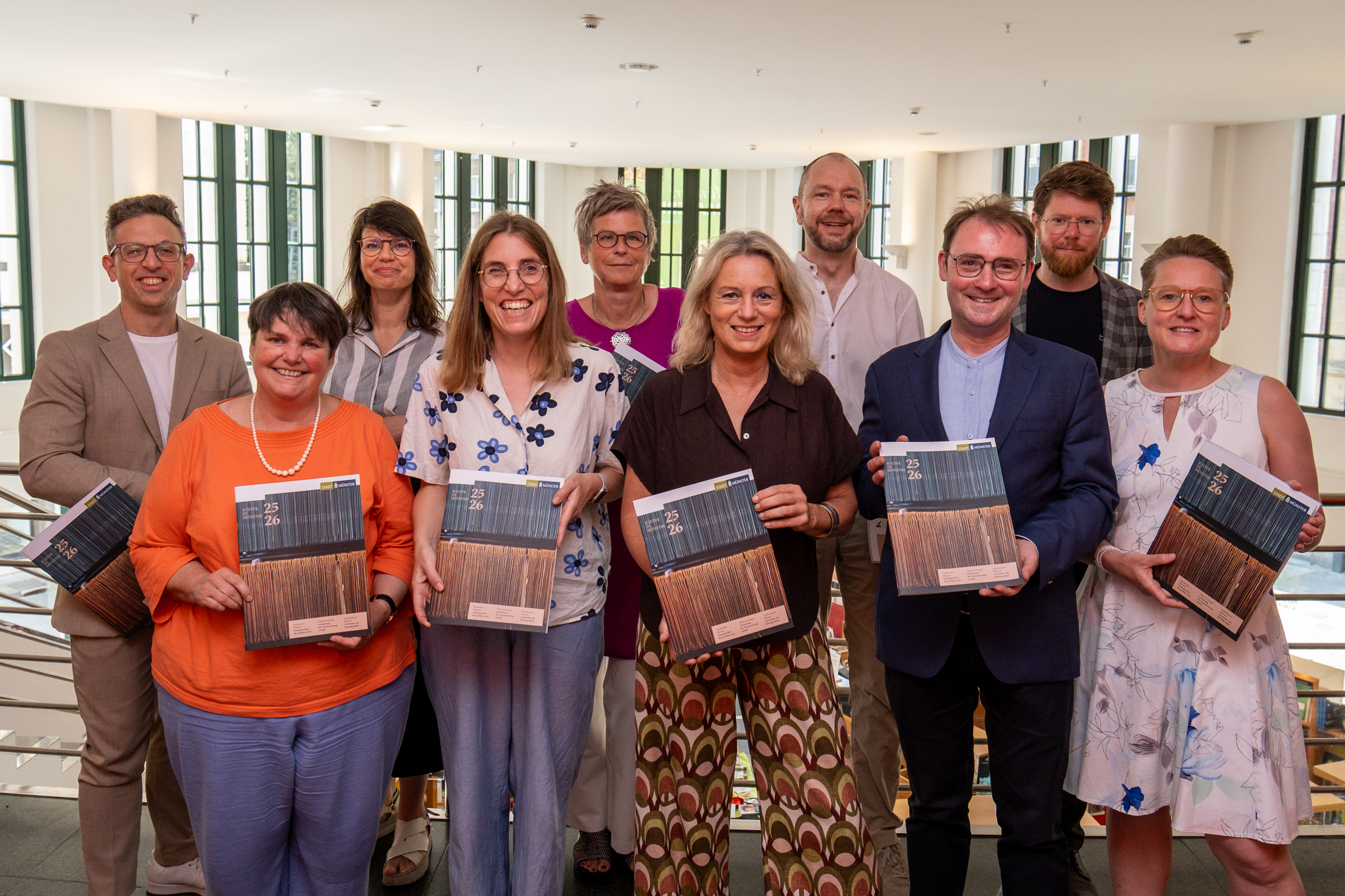 Zehn Personen stehen aufgereiht für ein Gruppenfoto; in der ersten Reihe hält man eine Zeitschrift in die Kamera.