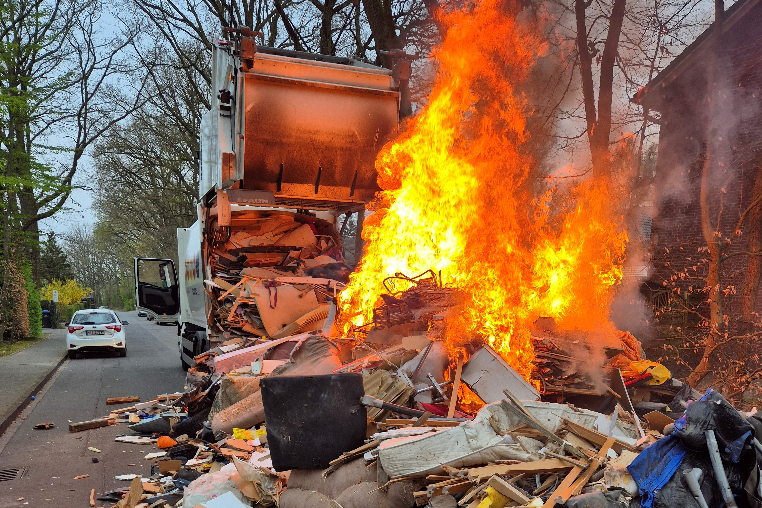 Ein in Flammen stehender Sperrmüllhaufen, ausgeschüttet auf der Straße. Dahinter das Sperrgutfahrzeug mit geöffneter Heckklappe.