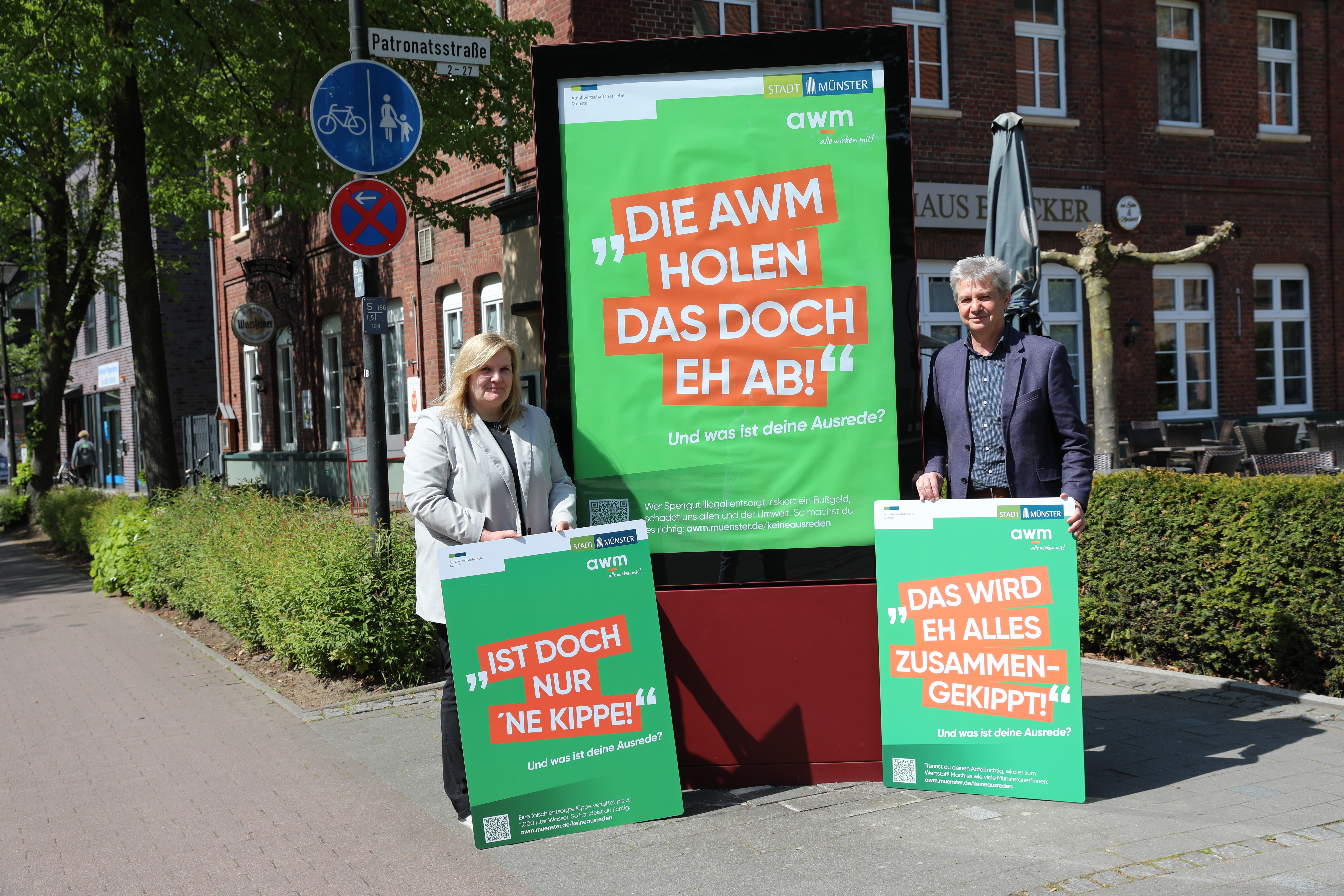 Christian Wedding und Manuela Feldkamp-Bode präsentieren die drei Kampagnenmotive. Jeweils eines halten sie in den Händen, das dritte hängt in einer Plakatwand in der Stadt, neben der die beiden Personen stehen.