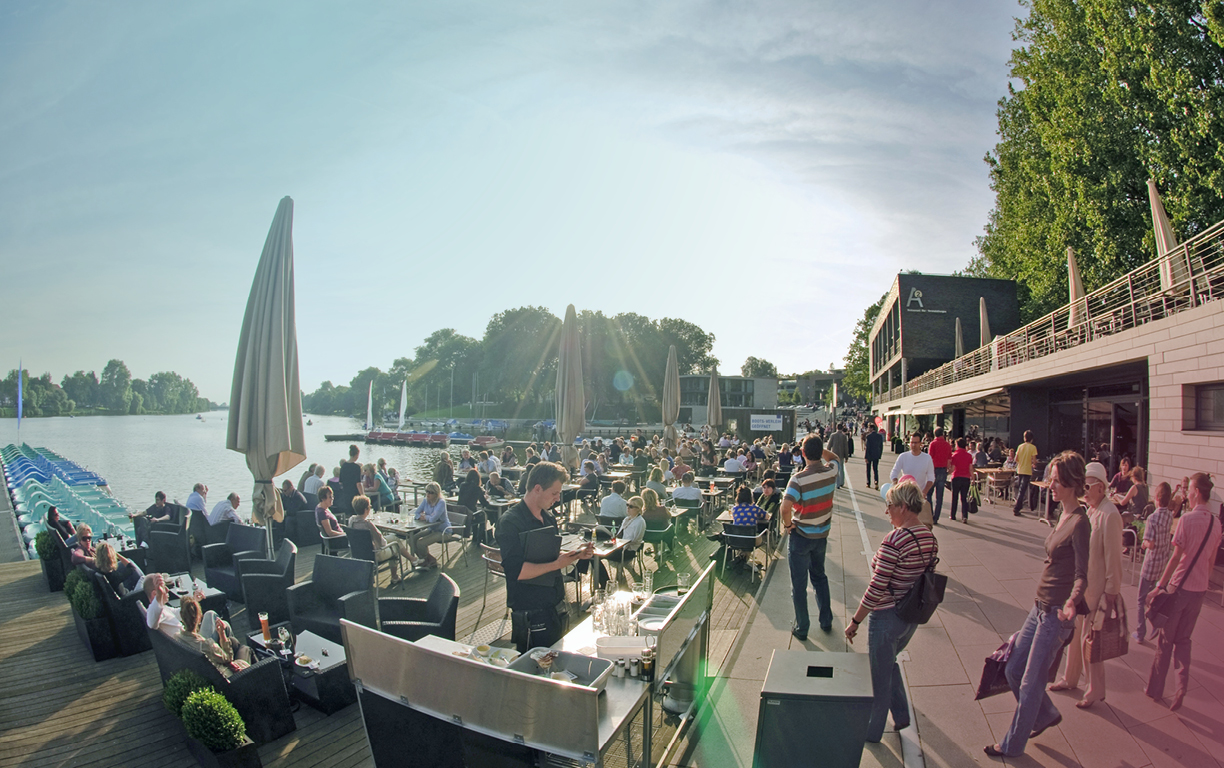 Zahlreiche Menschen sitzen an Tischen und Stühlen auf den Aaseeterrassen in Münster und die Sonne geht langsam hinter den Bäumen neben dem See unter.