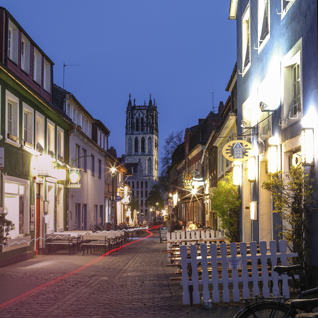 Eine kopfsteingepflasterte Straße mit Außengastronomie und beleuchteten Schildern der Restaurants in Münster mit Blick auf die Überwasserkirche