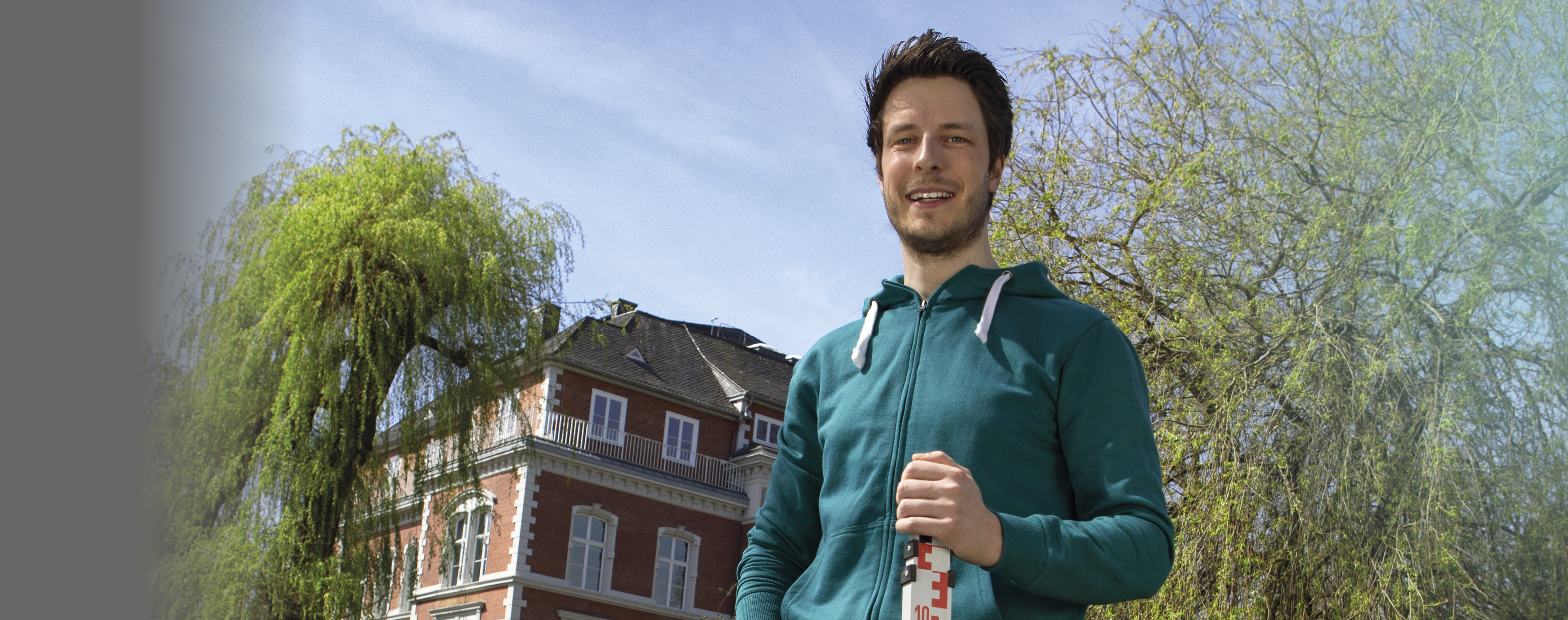 Ein junger Mann mit einer grünen Sweatshirtjacke steht vor der Musikschule Münster und hält einen Messstab für seine Arbeit als Ingenieur für Wasserwirtschaft