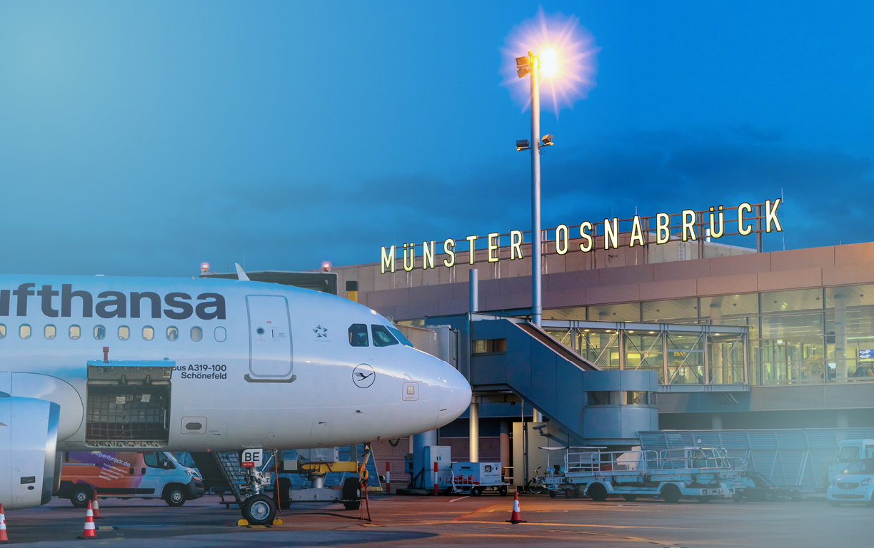 Ein Flugzeug der Lufthansa steht in der Abenddämmerung am Terminal des Flughafen Münster-Osnabrücks und wird von einem hellen Scheinwerfer angestrahlt.