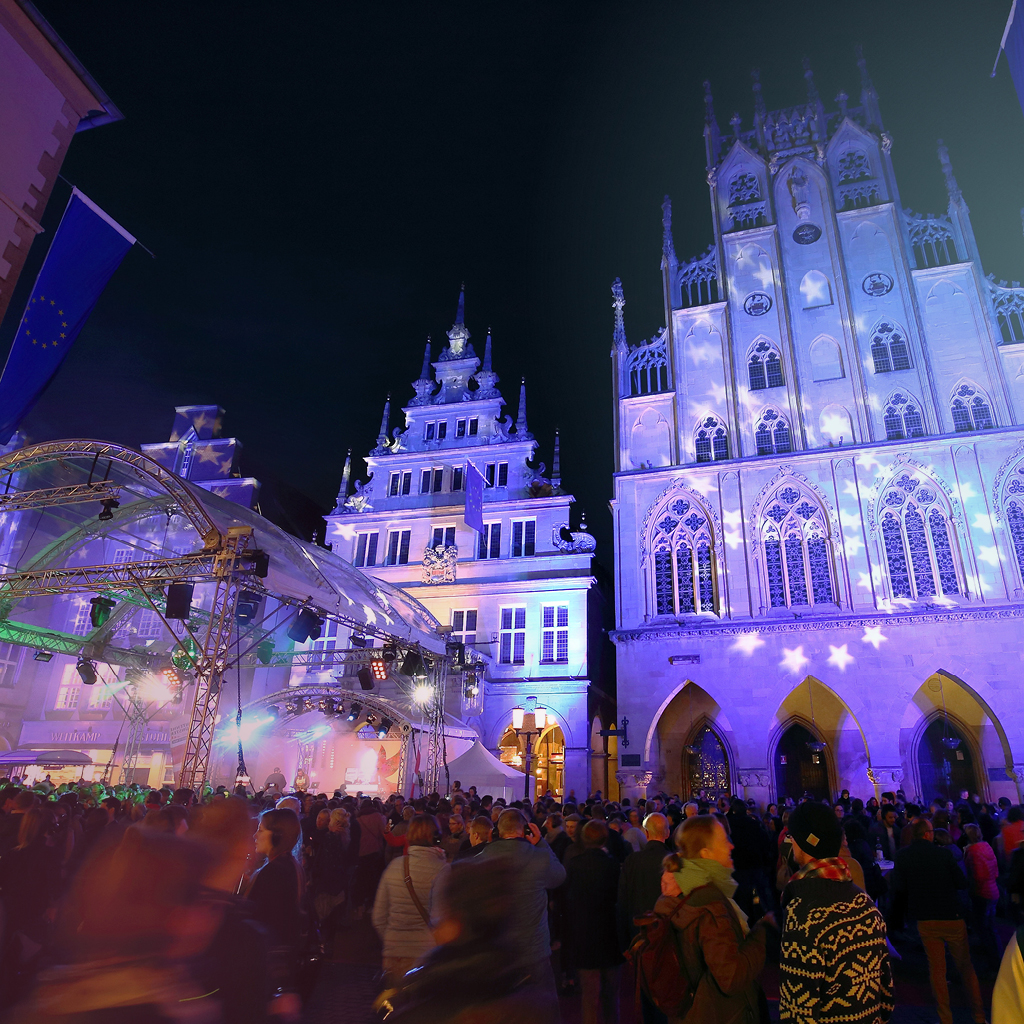 Eine Bühne ist auf dem Prinzipalmarkt aufgebaut und eine Menschenmenge feiert vor dem lila angeleuchteten Rathaus in Münster.