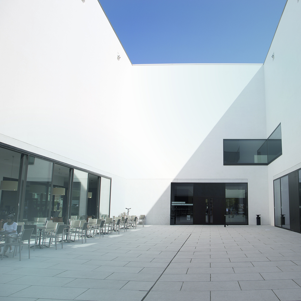 Gradlinige Architektur bildet sich in Form von weißen Wänden vor blauem Himmel ab und zeigt den Innenhof mit Eingang des LWL-Museums in Münster.