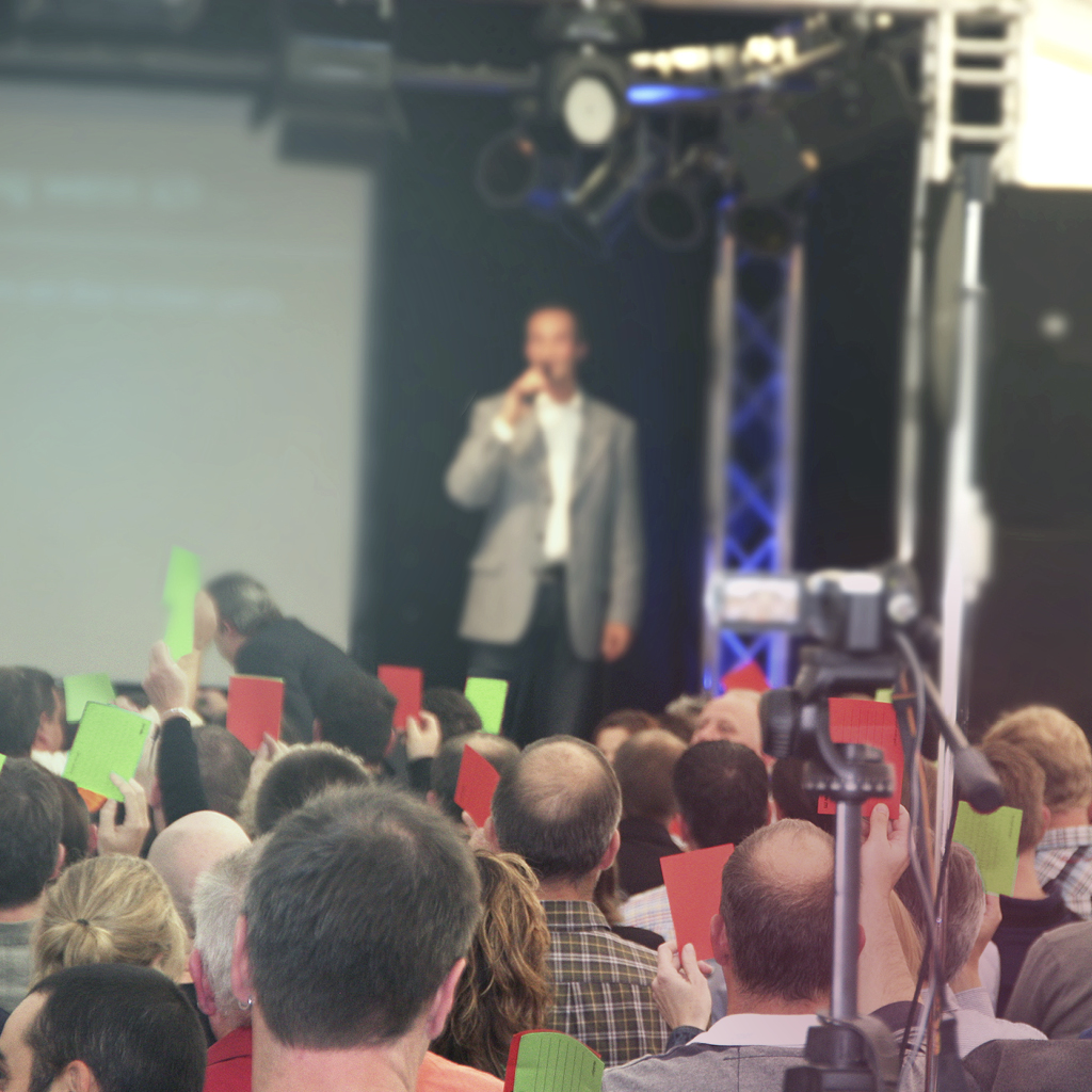 Eine große Gruppe von Menschen sitzt vor einer Bühne und hält rote und grüne Fähnchen in die Höhe, im Hintergrund steht ein Mann auf der Bühne und spricht in ein Mikrofon.