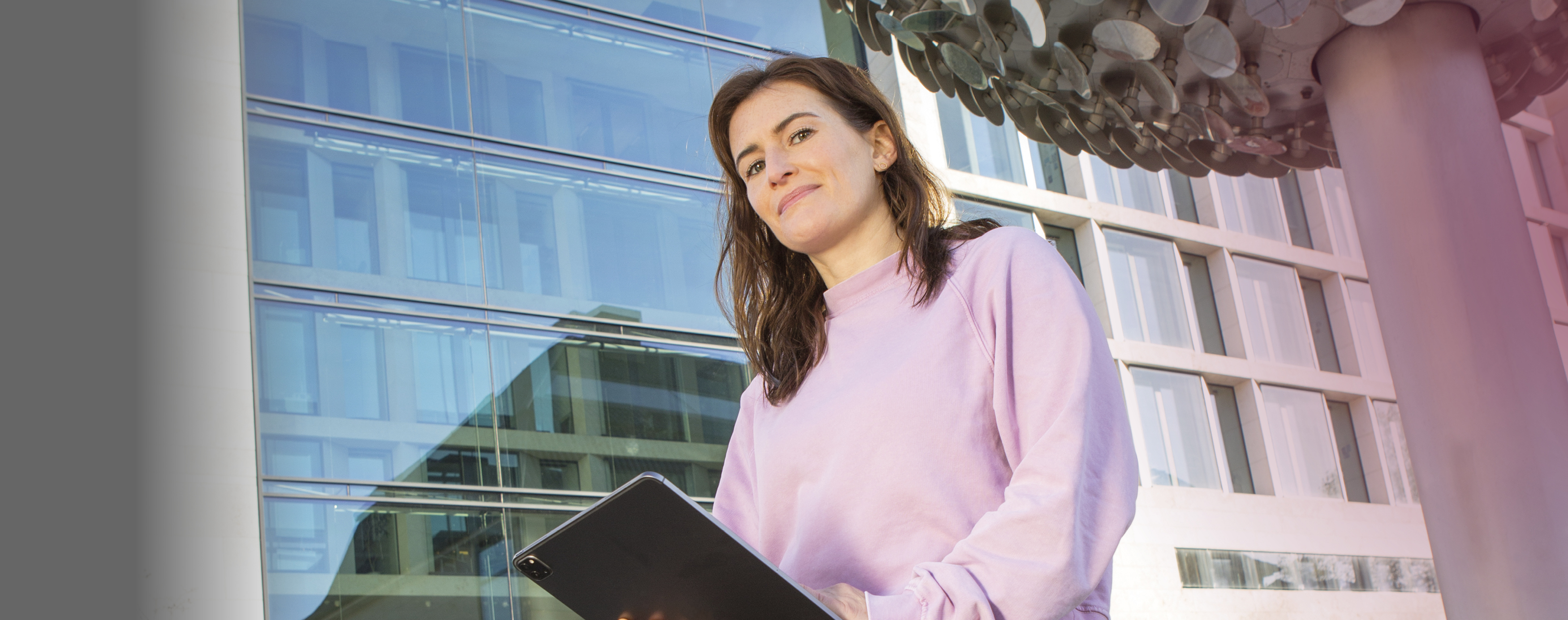 Ein Frau mit einem rosa Sweatshirtjacke sitzt auf einem Geländer vor der NRW-Bank und arbeitet mit ihrem Tablet von dort aud mobil.